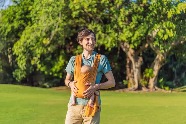 Babam küçük bebeğini parkta sarı bir askıda tutuyor. Sıcak ve sevgi dolu bir aile anı. Bebek giyimi, babalık ve ebeveyn-çocuk bağlama kavramı..
