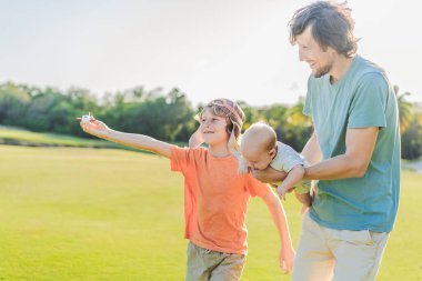 Baba ve iki oğlu parkta, büyük çocuk pilot şapkası takıyor ve bebek izlerken oyuncak bir uçak tutuyor. Çocukluk, bağ ve aile macerası konsepti.