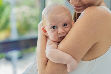 Anne evde bebeğine nazikçe sarılıyor, sıcak ve sevgi dolu bir anı paylaşıyor. Ebeveynlik, aile bağı ve anne sevgisi kavramı..