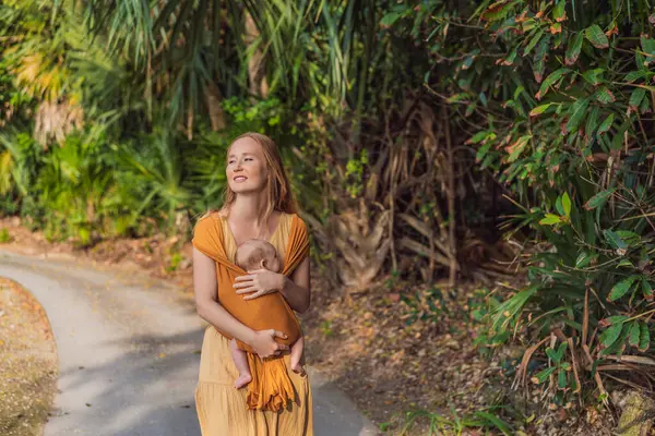 Anne, küçük bebeğini parkta sarı bir askıda tutuyor. Sıcak ve sevgi dolu bir aile anı. Bebek giyimi, ebeveynlik ve anne-çocuk bağlama kavramı.