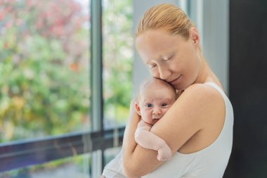 Anne evde bebeğine nazikçe sarılıyor, sıcak ve sevgi dolu bir anı paylaşıyor. Ebeveynlik, aile bağı ve anne sevgisi kavramı..