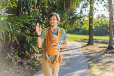 Babam küçük bebeğini parkta sarı bir askıda tutuyor. Sıcak ve sevgi dolu bir aile anı. Bebek giyimi, babalık ve ebeveyn-çocuk bağlama kavramı..