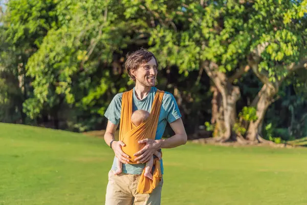 Babam küçük bebeğini parkta sarı bir askıda tutuyor. Sıcak ve sevgi dolu bir aile anı. Bebek giyimi, babalık ve ebeveyn-çocuk bağlama kavramı..