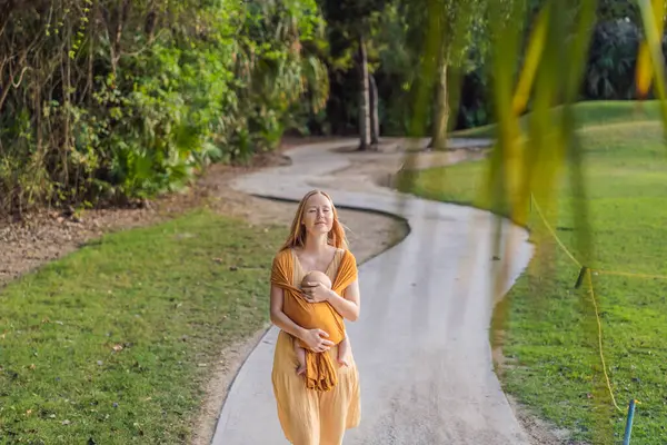 Anne, küçük bebeğini parkta sarı bir askıda tutuyor. Sıcak ve sevgi dolu bir aile anı. Bebek giyimi, ebeveynlik ve anne-çocuk bağlama kavramı.