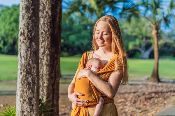 Anne, küçük bebeğini parkta sarı bir askıda tutuyor. Sıcak ve sevgi dolu bir aile anı. Bebek giyimi, ebeveynlik ve anne-çocuk bağlama kavramı.