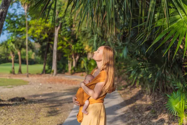 Anne, küçük bebeğini parkta sarı bir askıda tutuyor. Sıcak ve sevgi dolu bir aile anı. Bebek giyimi, ebeveynlik ve anne-çocuk bağlama kavramı.