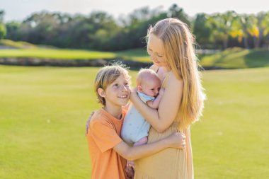 Anne, büyük oğul ve parkta bir çayırda yeni doğmuş bebek. Aile, doğada birlikte kaliteli zaman geçirir. Ebeveynlik, sevgi ve aile bağı kavramı.