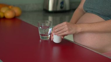 Parlak bir mutfakta duran hamile bir kadın, bir bardak suyla doğum öncesi vitamin hapları içiyor. Sağlığını ve bebeğinin gelişimini gözetiyor..