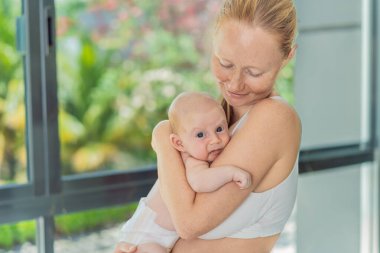 Anne evde bebeğine nazikçe sarılıyor, sıcak ve sevgi dolu bir anı paylaşıyor. Ebeveynlik, aile bağı ve anne sevgisi kavramı..