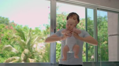 Genç baba, arka planda tropik manzarası olan büyük bir pencerenin yanında 3 aylık oğluyla egzersiz yapıyor..