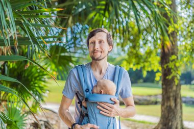 Genç bir baba, bebeğiyle birlikte doğada ebeveynlere bakan ergonomik bir bebek taşıyıcısının içinde dolaşıyor. Barışçıl bir ortamda bağlılık, sıcaklık ve modern ebeveynlik kavramı.