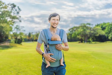 Genç bir baba, bebeğiyle birlikte doğada ebeveynlere bakan ergonomik bir bebek taşıyıcısının içinde dolaşıyor. Barışçıl bir ortamda bağlılık, sıcaklık ve modern ebeveynlik kavramı.