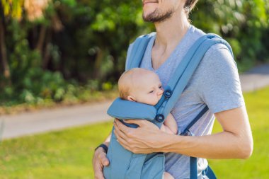 Genç bir baba, bebeğiyle birlikte doğada ebeveynlere bakan ergonomik bir bebek taşıyıcısının içinde dolaşıyor. Barışçıl bir ortamda bağlılık, sıcaklık ve modern ebeveynlik kavramı.
