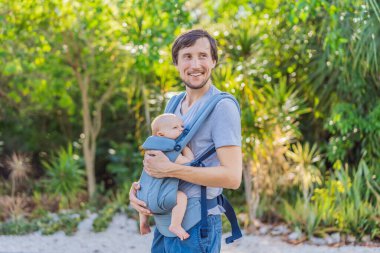 Genç bir baba, bebeğiyle birlikte doğada ebeveynlere bakan ergonomik bir bebek taşıyıcısının içinde dolaşıyor. Barışçıl bir ortamda bağlılık, sıcaklık ve modern ebeveynlik kavramı.