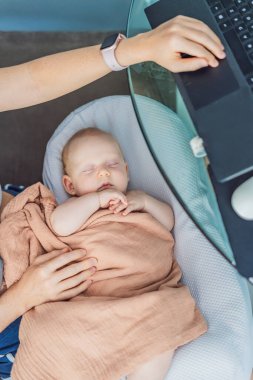 Yeni doğmuş bebeği kucağında rahat bir bebek yuvasında huzur içinde uyurken aynı işi yapan anne evde dizüstü bilgisayarla çalışıyor. Uzaktan çalışma, annelik, aile ve kariyer dengeleme kavramı.