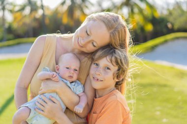 Anne, büyük oğul ve parkta bir çayırda yeni doğmuş bebek. Aile, doğada birlikte kaliteli zaman geçirir. Ebeveynlik, sevgi ve aile bağı kavramı.