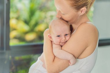 Anne evde bebeğine nazikçe sarılıyor, sıcak ve sevgi dolu bir anı paylaşıyor. Ebeveynlik, aile bağı ve anne sevgisi kavramı..
