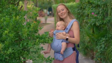 Genç anne, güneşli bir günde bebeğiyle parka gidip ergonomik bir taşıyıcıyla kaliteli zaman ve doğanın tadını çıkarıyor..