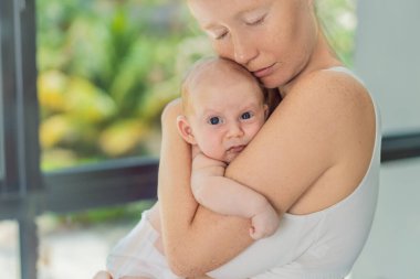 Anne evde bebeğine nazikçe sarılıyor, sıcak ve sevgi dolu bir anı paylaşıyor. Ebeveynlik, aile bağı ve anne sevgisi kavramı..