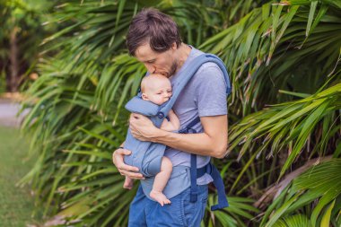Genç bir baba, bebeğiyle birlikte doğada ebeveynlere bakan ergonomik bir bebek taşıyıcısının içinde dolaşıyor. Barışçıl bir ortamda bağlılık, sıcaklık ve modern ebeveynlik kavramı.
