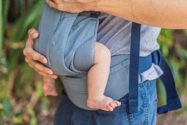 Genç bir baba, bebeğiyle birlikte doğada ebeveynlere bakan ergonomik bir bebek taşıyıcısının içinde dolaşıyor. Barışçıl bir ortamda bağlılık, sıcaklık ve modern ebeveynlik kavramı.