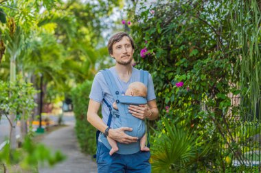 Genç bir baba, bebeğiyle birlikte doğada ebeveynlere bakan ergonomik bir bebek taşıyıcısının içinde dolaşıyor. Barışçıl bir ortamda bağlılık, sıcaklık ve modern ebeveynlik kavramı.