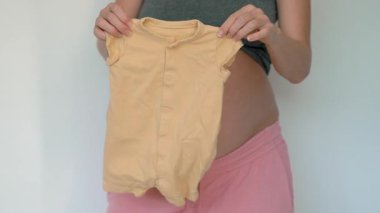 Hamile kadın yeni doğmuş bebek kıyafetlerini karnının yanında tutuyor. Çocuklarına hazırlanırken heyecanını ve sevgisini gösteriyor..