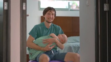 Bir baba kundaklanmış yeni doğmuş bebeğini bir fitbol topunun üzerinde otururken nazikçe sallayarak uyutur. Bebek sakin ve rahatlamış. Huzurlu bir ebeveyn, bağ kurma ve yatma zamanı bakımı sahnesi.