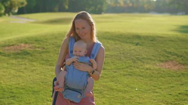 Genç anne, güneşli bir günde bebeğiyle parka gidip ergonomik bir taşıyıcıyla kaliteli zaman ve doğanın tadını çıkarıyor..