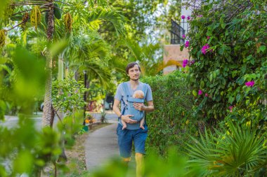 Genç bir baba, bebeğiyle birlikte doğada ebeveynlere bakan ergonomik bir bebek taşıyıcısının içinde dolaşıyor. Barışçıl bir ortamda bağlılık, sıcaklık ve modern ebeveynlik kavramı.