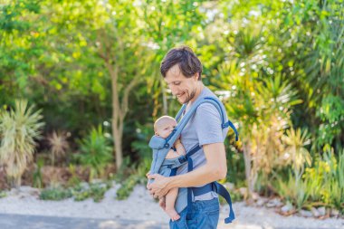 Genç bir baba, bebeğiyle birlikte doğada ebeveynlere bakan ergonomik bir bebek taşıyıcısının içinde dolaşıyor. Barışçıl bir ortamda bağlılık, sıcaklık ve modern ebeveynlik kavramı.