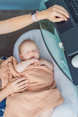 Yeni doğmuş bebeği kucağında rahat bir bebek yuvasında huzur içinde uyurken aynı işi yapan anne evde dizüstü bilgisayarla çalışıyor. Uzaktan çalışma, annelik, aile ve kariyer dengeleme kavramı.