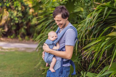 Genç bir baba, bebeğiyle birlikte doğada ebeveynlere bakan ergonomik bir bebek taşıyıcısının içinde dolaşıyor. Barışçıl bir ortamda bağlılık, sıcaklık ve modern ebeveynlik kavramı.