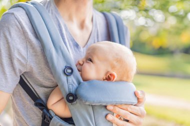 Genç bir baba, bebeğiyle birlikte doğada ebeveynlere bakan ergonomik bir bebek taşıyıcısının içinde dolaşıyor. Barışçıl bir ortamda bağlılık, sıcaklık ve modern ebeveynlik kavramı.