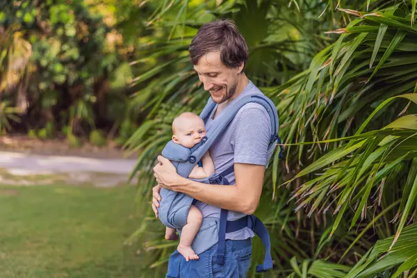 Genç bir baba, bebeğiyle birlikte doğada ebeveynlere bakan ergonomik bir bebek taşıyıcısının içinde dolaşıyor. Barışçıl bir ortamda bağlılık, sıcaklık ve modern ebeveynlik kavramı.