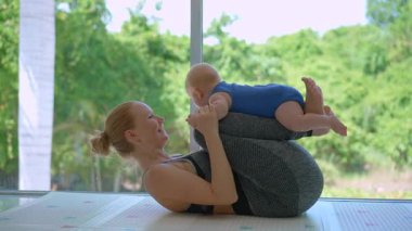 Genç anne, arka planda tropik manzarası olan büyük bir pencerenin yanında 3 aylık oğluyla egzersiz yapıyor..