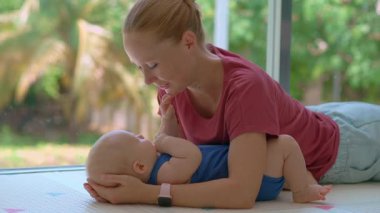 Annem 3 aylık bebeğini tropikal arka planı olan büyük bir pencerenin yanında öpüyor. Şefkat bağı ve erken annelik kavramı.