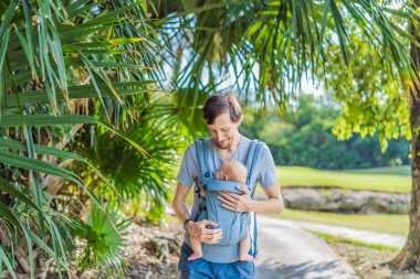 Genç bir baba, bebeğiyle birlikte doğada ebeveynlere bakan ergonomik bir bebek taşıyıcısının içinde dolaşıyor. Barışçıl bir ortamda bağlılık, sıcaklık ve modern ebeveynlik kavramı.