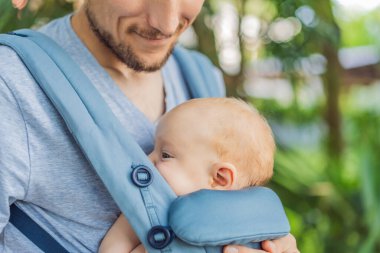 Genç bir baba, bebeğiyle birlikte doğada ebeveynlere bakan ergonomik bir bebek taşıyıcısının içinde dolaşıyor. Barışçıl bir ortamda bağlılık, sıcaklık ve modern ebeveynlik kavramı.