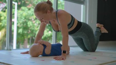 Genç anne, arka planda tropik manzarası olan büyük bir pencerenin yanında 3 aylık oğluyla egzersiz yapıyor..