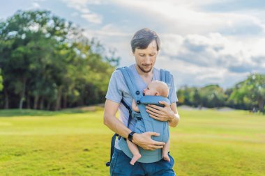 Genç bir baba, bebeğiyle birlikte doğada ebeveynlere bakan ergonomik bir bebek taşıyıcısının içinde dolaşıyor. Barışçıl bir ortamda bağlılık, sıcaklık ve modern ebeveynlik kavramı.