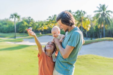 Baba ve iki oğlu parkta, büyük çocuk pilot şapkası takıyor ve bebek izlerken oyuncak bir uçak tutuyor. Çocukluk, bağ ve aile macerası konsepti.