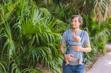 Genç bir baba, bebeğiyle birlikte doğada ebeveynlere bakan ergonomik bir bebek taşıyıcısının içinde dolaşıyor. Barışçıl bir ortamda bağlılık, sıcaklık ve modern ebeveynlik kavramı.