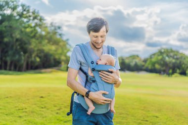 Genç bir baba, bebeğiyle birlikte doğada ebeveynlere bakan ergonomik bir bebek taşıyıcısının içinde dolaşıyor. Barışçıl bir ortamda bağlılık, sıcaklık ve modern ebeveynlik kavramı.