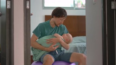 Bir baba kundaklanmış yeni doğmuş bebeğini bir fitbol topunun üzerinde otururken nazikçe sallayarak uyutur. Bebek sakin ve rahatlamış. Huzurlu bir ebeveyn, bağ kurma ve yatma zamanı bakımı sahnesi.