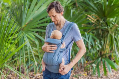 Genç bir baba, bebeğiyle birlikte doğada ebeveynlere bakan ergonomik bir bebek taşıyıcısının içinde dolaşıyor. Barışçıl bir ortamda bağlılık, sıcaklık ve modern ebeveynlik kavramı.