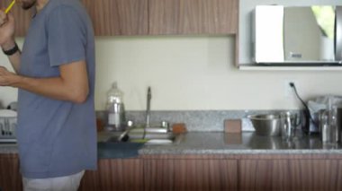 Mutfakta dişlerini fırçalarken telefonunu kullanan genç bir adam, birden fazla iş yapmanın ve telefon bağımlılığının modern bir karışımını sergiliyor. Teknolojinin günlük hayattaki rolünü yansıtan bir an.