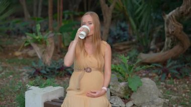 Hamile bir kadın tropik bir parkta bankta otururken kahve içiyor. Hamilelik boyunca sağlık sorunları ve kafein tüketimi riskleri kavramı.