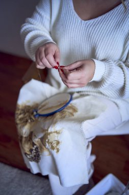               Hafif giysili orta boy bir kadın, beyaz bir odada beyaz bir sandalyede otururken altın ve bej renkte bir resim nakışlıyor.                