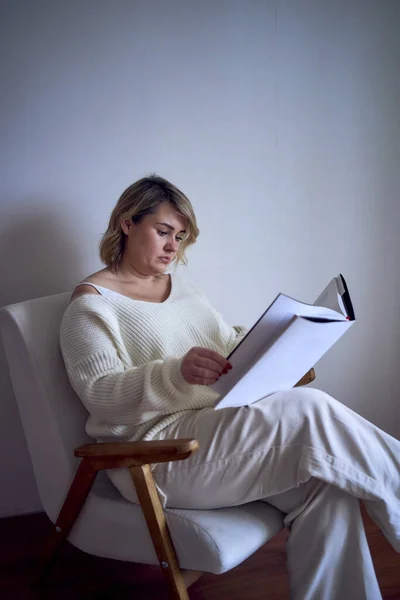 medium-sized woman in light clothes reads a book while sitting in a white chair in a light room